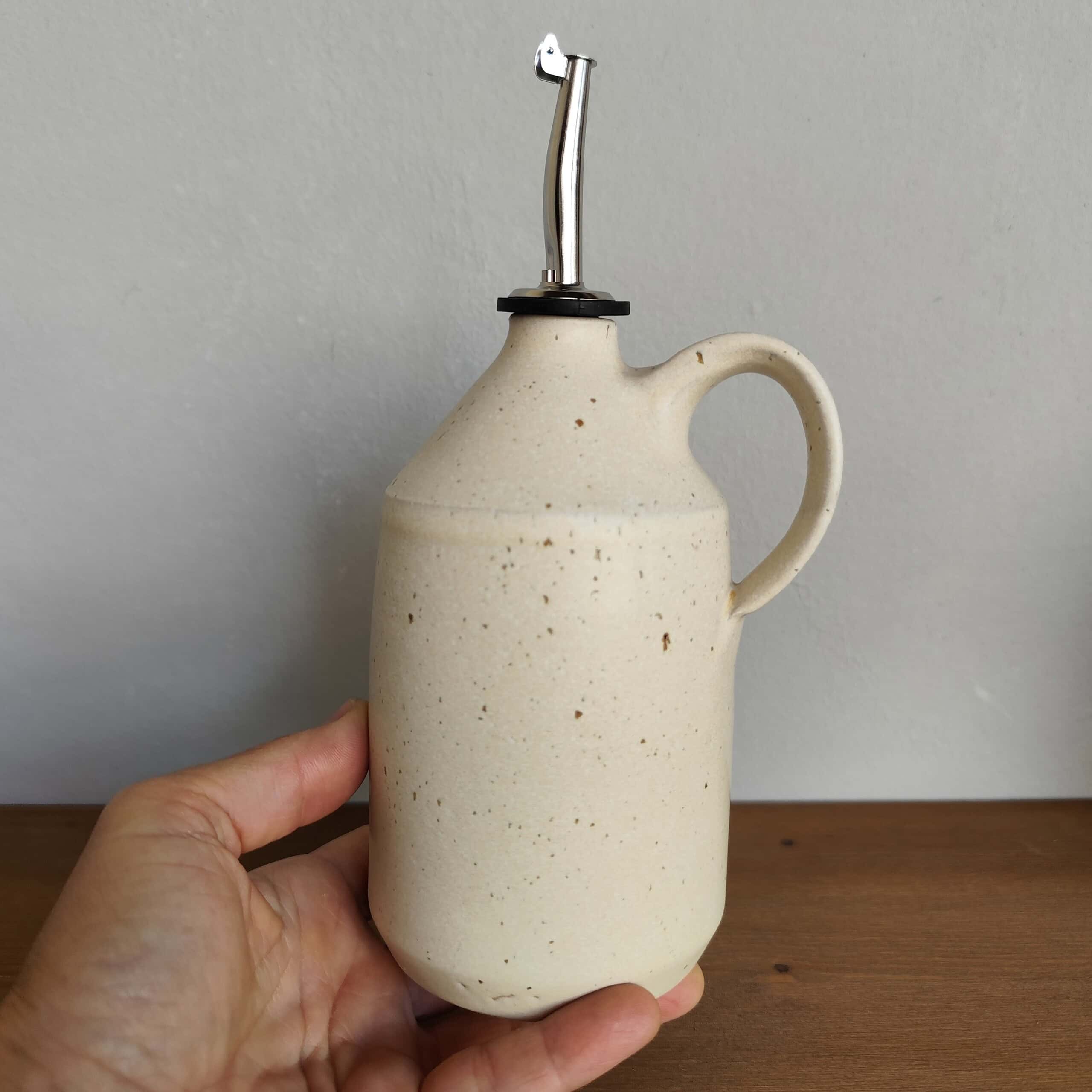 Oliera avorio in gres con manico, smalto avorio satinato e beccuccio in acciaio inox sostenuta da una mano