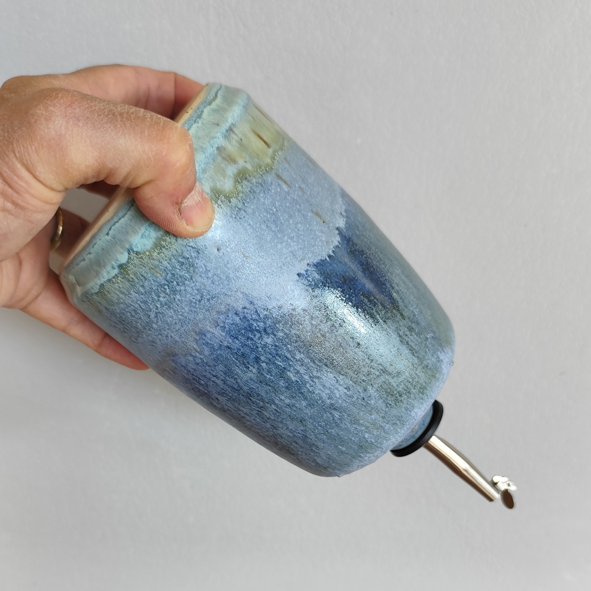 oliera blu variegato in gres con smalti sovrapposti, zone verde acqua e turchese tenuta in mano come se stesse versando tramite il beccuccio in acciaio inox